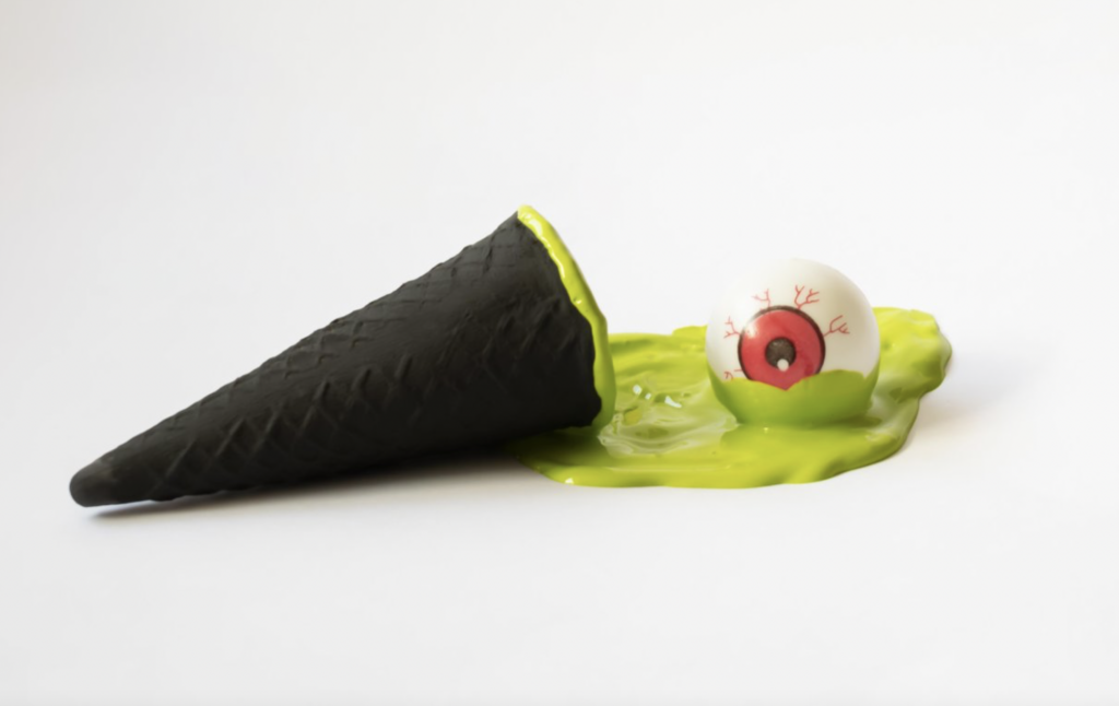 Conceptual photo of a black ice cream cone with green, slimy ice cream melting on a white table and a fake eyeball as a Halloween decoration