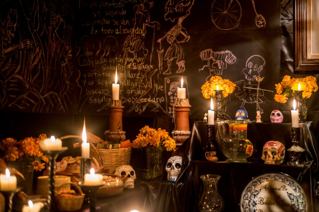 Moody and dark photo of a Dia de los Muertos (day of the dead) ofrenda altar with painted calavera sugar skulls and glowing lit candles against a chalk board